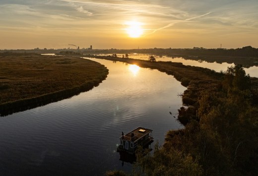 Hausboot auf der Peene bei Sonnenuntergang, umgeben von ruhiger Flusslandschaft und goldenem Licht am Horizont.