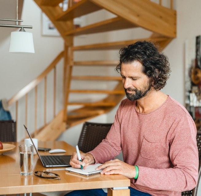 Ein Mann sitzt an einem Holztisch in einem Ferienhaus, arbeitet am Laptop und macht Notizen – ideale Bedingungen für eine Workation auf der Insel Poel.