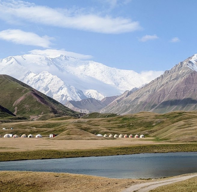 Ein Bild von der Landschaft Kirgistan. Im Hintergrund ist ein Gebirgsmantel, bestehend aus dreu hohen, schneebedeckten Bergen zu sehen.
Davor stehen Hütten von den dortlebenden Menschen und ein Gewässer befindet sich vor ihnen., © Katrin Fischbeck