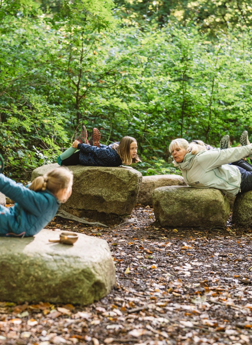 Kinder und ein Erwachsener liegen auf großen Steinen im Wald und machen gemeinsam Yoga-Übungen, umgeben von dichten Bäumen und Blättern.