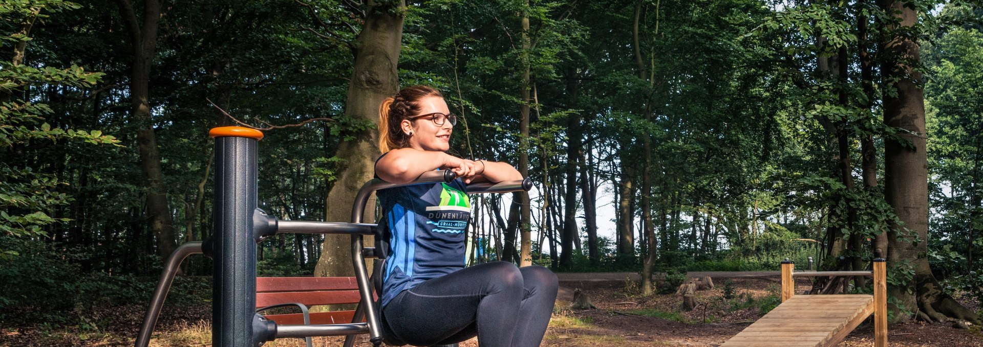 Eine Frau trainiert an einem Fitnessgerät im Aktivwald Graal-Müritz, umgeben von hohen Bäumen und natürlicher Waldatmosphäre.