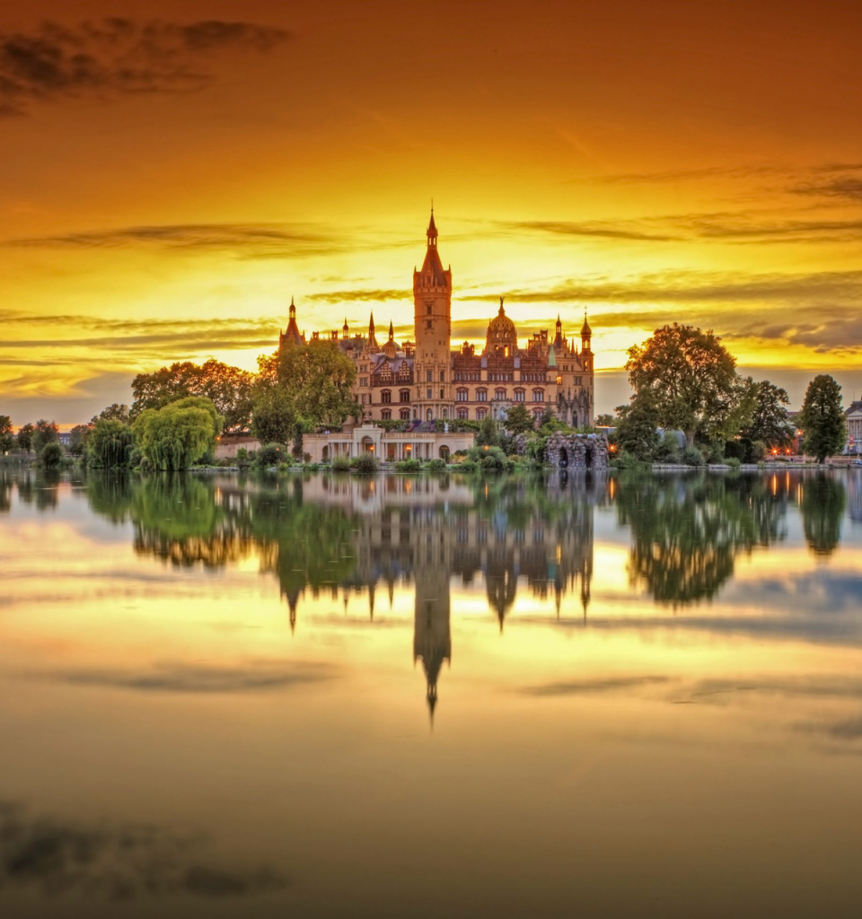 Das Schweriner Schloss wird von den letzten Sonnenstrahlen umrahmt. Der Sonnenuntergang färbt den Himmel orange. Im ruhigen See spiegelt sich das Schweriner Schloss.