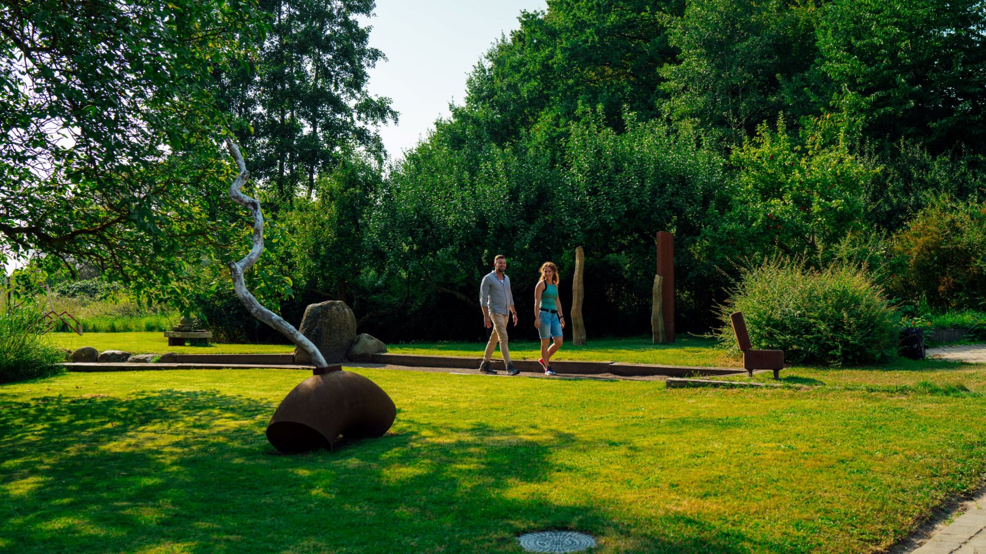 Ein Paar spaziert durch einen grünen Skulpturengarten in der Alten Wassermühle in Wreechen. Im Vordergrund steht eine moderne Kunstinstallation aus Metall, während Bäume und Büsche den Garten umrahmen. Die ruhige Atmosphäre lädt zum Erkunden und Verweilen ein.