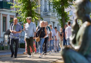 Eine Gruppe Menschen ist mit einem Stadtführer auf einem gepflasterten Weg unterwegs. Rechts, unscharf im Bild ist eine Statue am Pfaffenteich zu sehen, im Hintergrund erscheinen die Villen. Blickpunkt sind der Stadtführer sowie ein Paar, dass sich angeregt unterhält., © Oliver Borchert