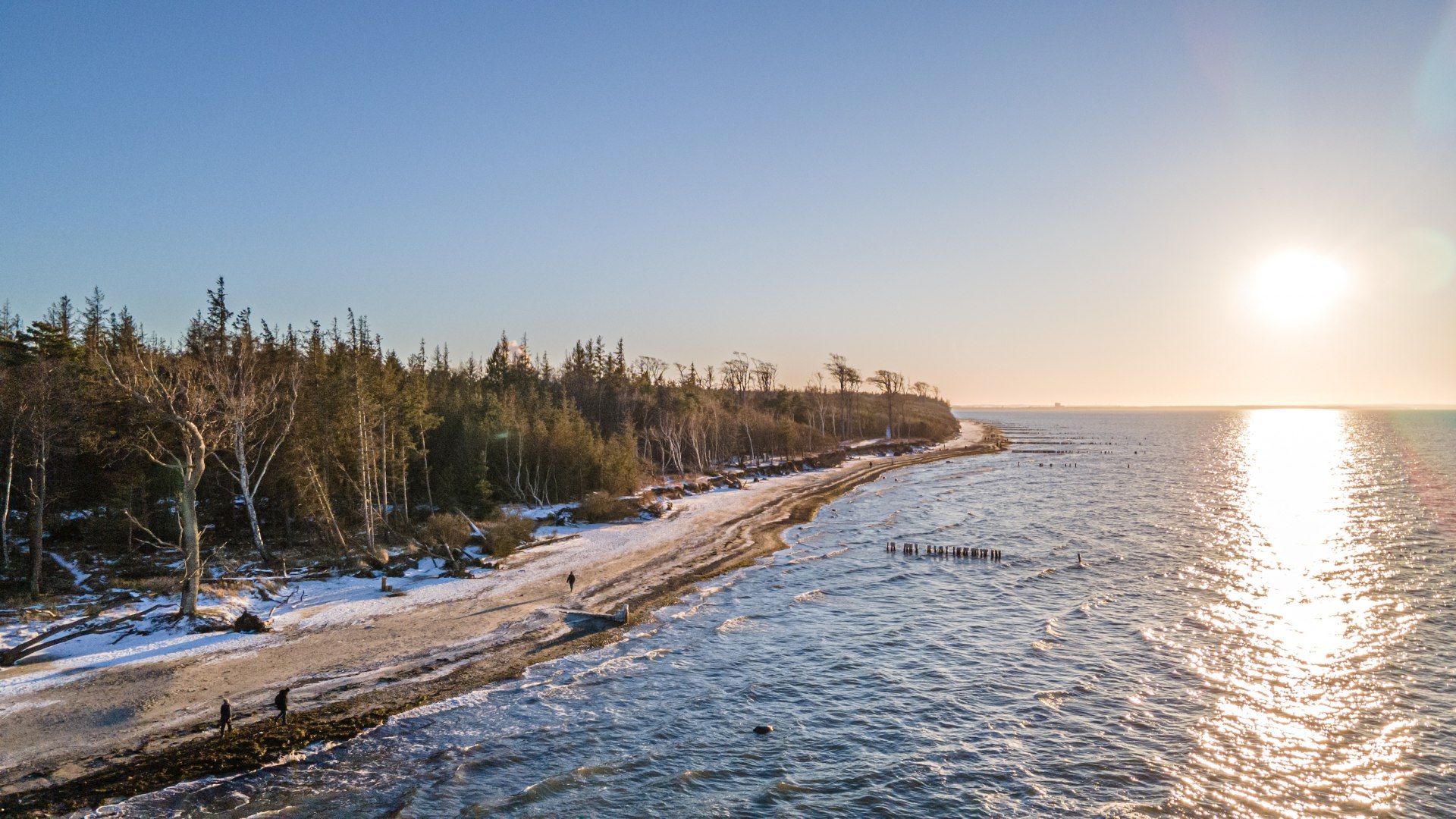 Winterlicher Strand bei der Torfbrücke mit schneebedeckten Bäumen, glitzernder Ostsee und einem Sonnenuntergang am Horizont.