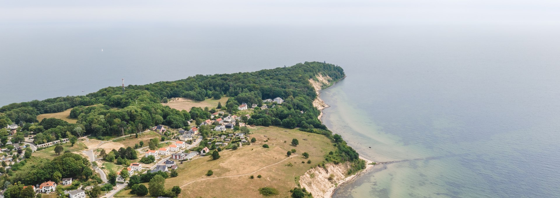 Luftaufnahme des Ostseebads Göhren auf Rügen mit weiten Wiesen, einer Steilküste und der Ostsee im Hintergrund unter leicht bewölktem Himmel.