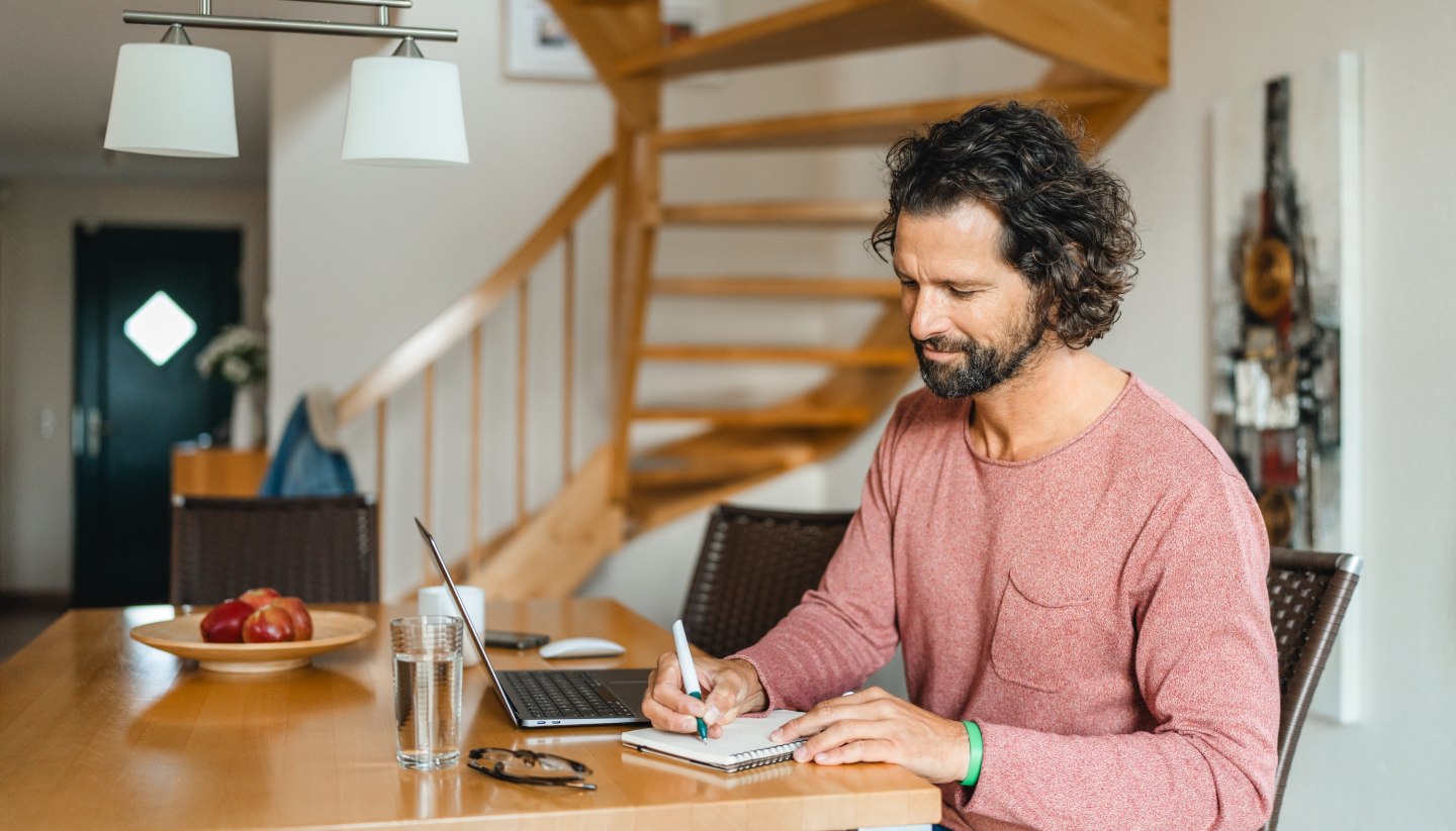 Ein Mann sitzt an einem Holztisch in einem Ferienhaus, arbeitet am Laptop und macht Notizen – ideale Bedingungen für eine Workation auf der Insel Poel.