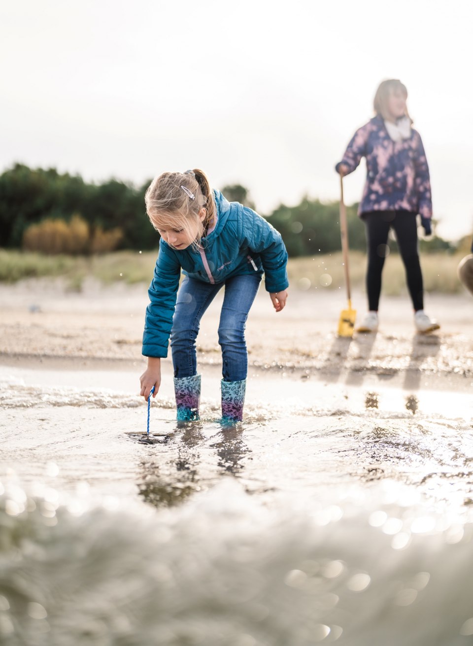 Ein Mädchen in Gummistiefeln steht gebückt im Wasser am Strand und sucht nach etwas im Sand, während ein weiteres Kind im Hintergrund steht.