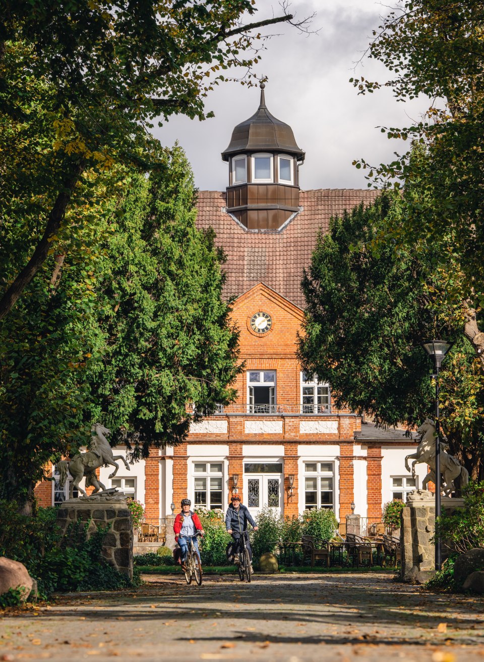 Zwei Radfahrer nähern sich dem Eingang des Schloss Basthorst, eingerahmt von Bäumen und herbstlicher Atmosphäre.