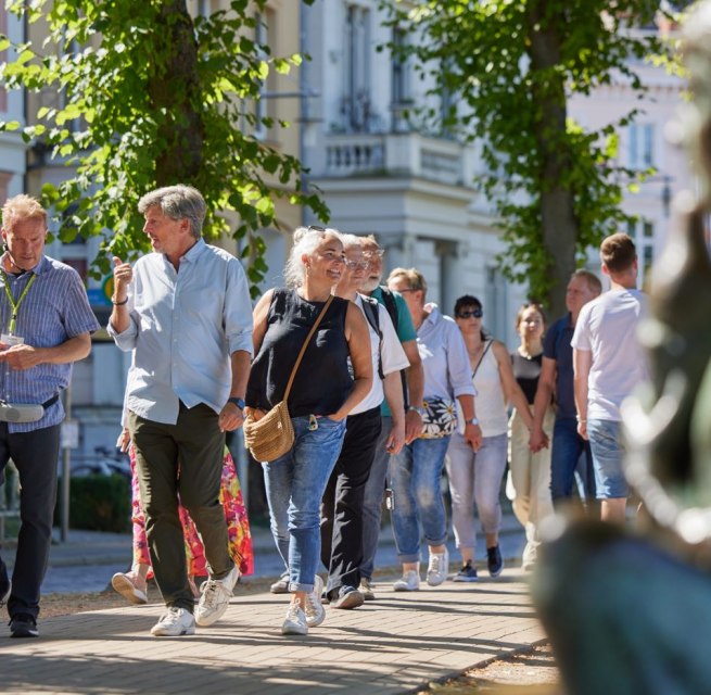 Eine Gruppe Menschen ist mit einem Stadtführer auf einem gepflasterten Weg unterwegs. Rechts, unscharf im Bild ist eine Statue am Pfaffenteich zu sehen, im Hintergrund erscheinen die Villen. Blickpunkt sind der Stadtführer sowie ein Paar, dass sich angeregt unterhält., © Oliver Borchert