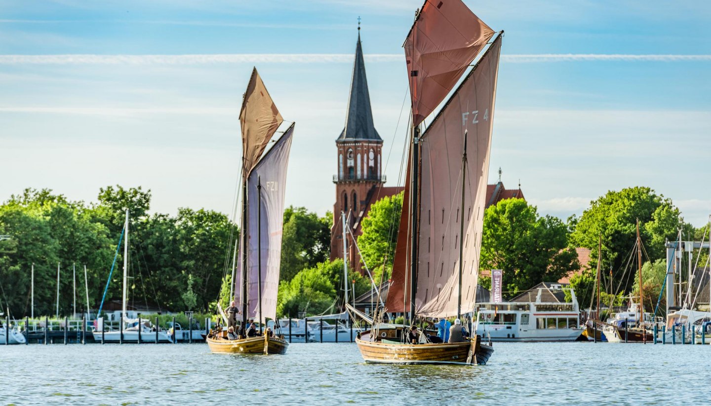 Zwei Zeesenboote segeln auf dem Bodden vor dem Hafen Wustrow mit Blick auf die Dorfkirche.