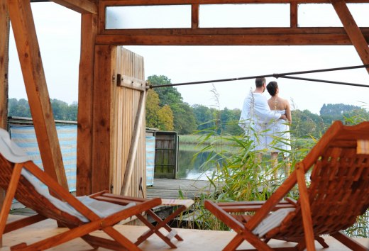 Ein Paar in Handtüchern steht auf einer Terrasse am See, umgeben von Schilf, mit Blick auf das Wasser in der Feldberger Seenlandschaft. Im Vordergrund sind Holzliegen zu sehen.