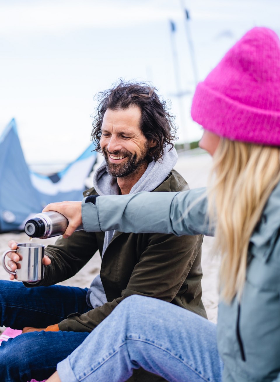 Ein Paar sitzt am Strand von Timmendorf, lächelt und genießt eine heiße Tasse Kaffee mit einem Kite im Hintergrund.