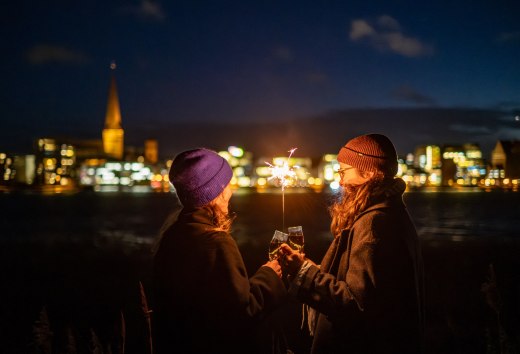 Zwei Personen stoßen mit Sekt an, während sie eine Wunderkerze halten, mit der beleuchteten Skyline von Rostock in Mecklenburg-Vorpommern im Hintergrund.