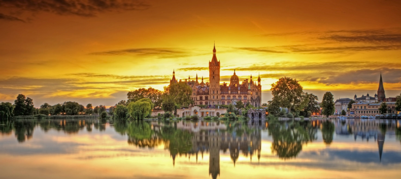 Das Schweriner Schloss wird von den letzten Sonnenstrahlen umrahmt. Der Sonnenuntergang färbt den Himmel orange. Im ruhigen See spiegelt sich das Schweriner Schloss.