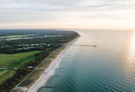 Luftaufnahme der Seebrücke und des Strandes von Graal-Müritz, umgeben von Wald und Wiesen, mit der Ostsee im Licht eines stimmungsvollen Sonnenuntergangs.