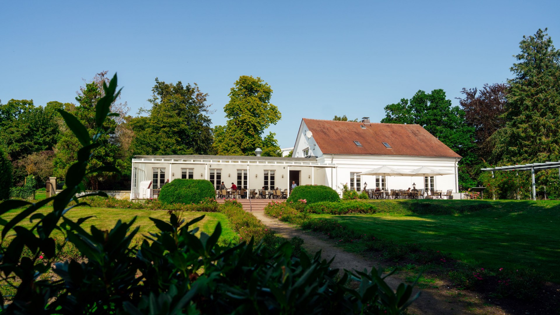 Das Rosencafé in Putbus auf Rügen, ein weißes Gebäude mit rotem Dach, umgeben von gepflegten Gärten und grünen Büschen. Vor dem Café laden Tische auf der Terrasse zu entspannten Stunden unter freiem Himmel ein. Im Hintergrund ragen hohe Bäume in den blauen Himmel.