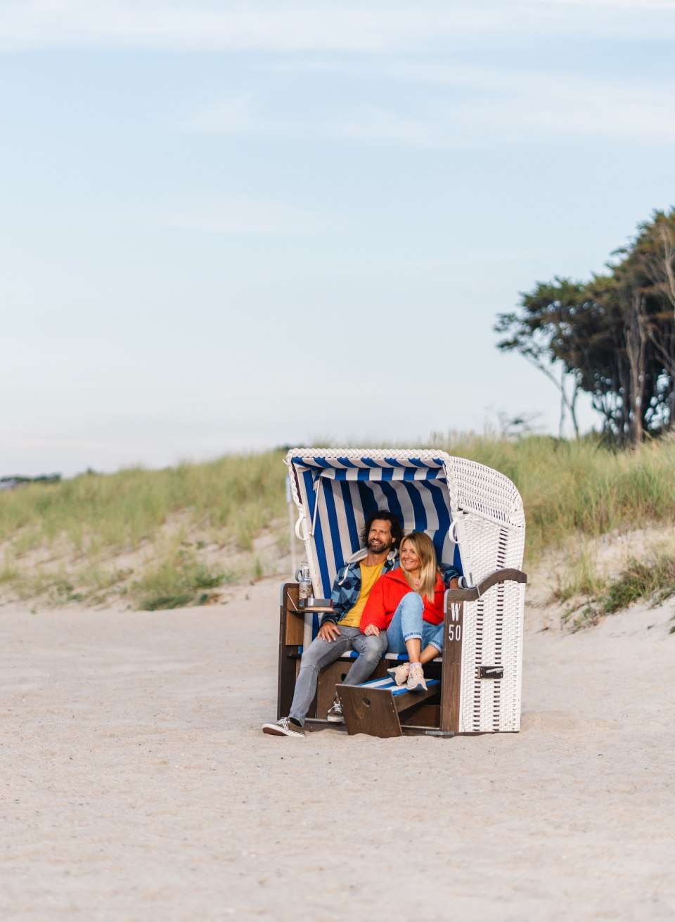 Ein Paar sitzt in einem blau-weiß gestreiften Strandkorb am Strand von Graal-Müritz, umgeben von Sand und Dünen mit Blick auf die ruhige Küstenlandschaft.