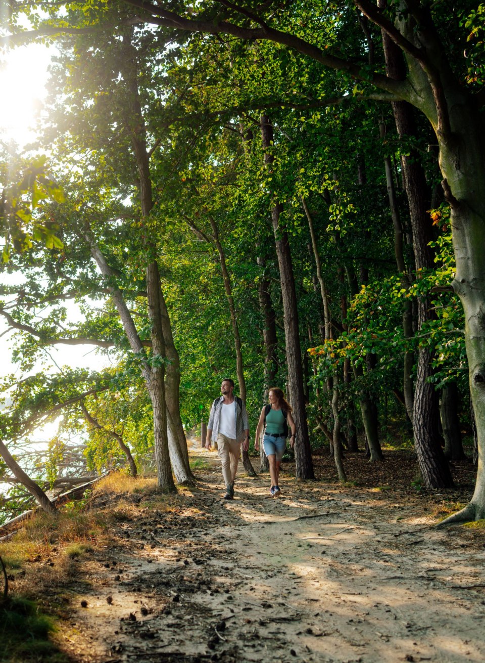 Ein Paar wandert auf einem schattigen Waldweg entlang der Küste im Biosphärenreservat Südost-Rügen. Die Sonne scheint durch die Bäume und spiegelt sich im Wasser. Die Atmosphäre ist ruhig und von der natürlichen Schönheit der Umgebung geprägt.