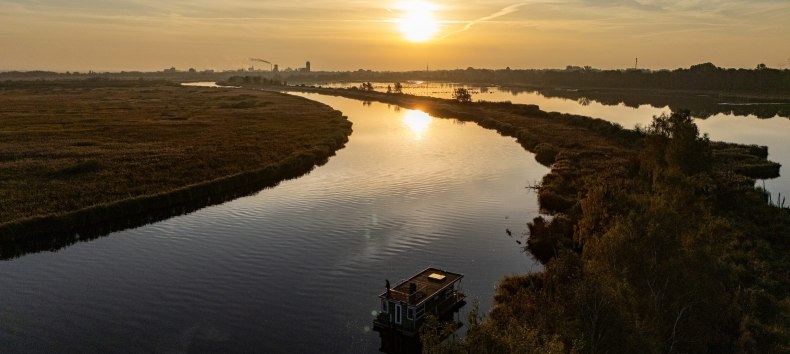 Hausboot auf der Peene bei Sonnenuntergang, umgeben von ruhiger Flusslandschaft und goldenem Licht am Horizont.