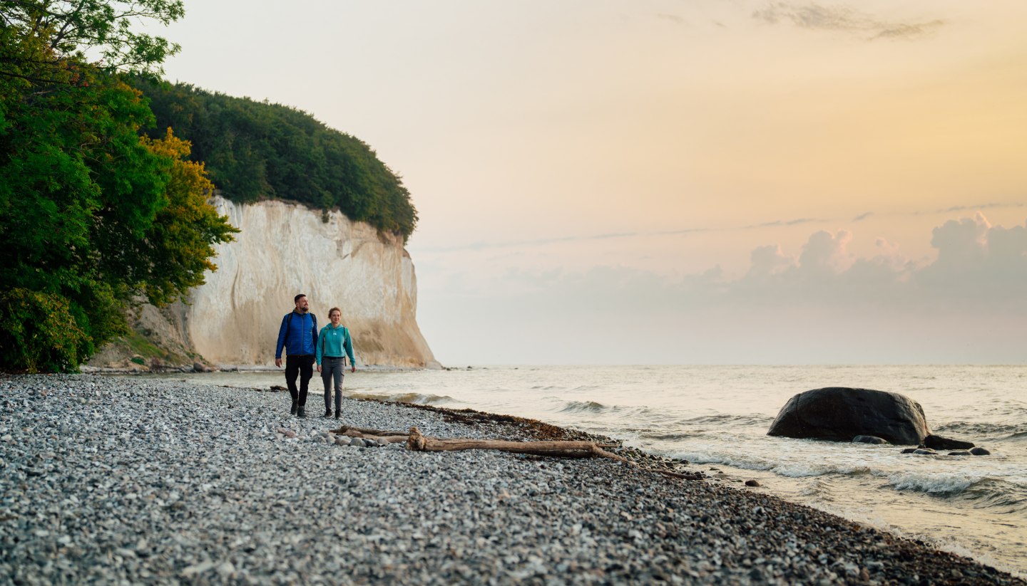 Ein Paar spaziert am Fuße der berühmten Kreidefelsen auf der Insel Rügen entlang, während der Sonnenuntergang den Himmel in sanften Farben erstrahlen lässt.