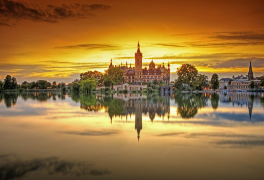 Das Schweriner Schloss wird von den letzten Sonnenstrahlen umrahmt. Der Sonnenuntergang färbt den Himmel orange. Im ruhigen See spiegelt sich das Schweriner Schloss.