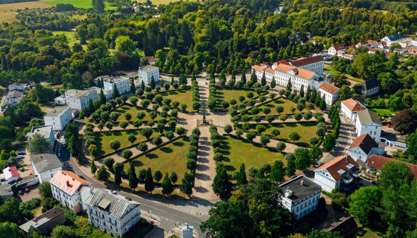 Luftaufnahme des Circus in Putbus auf Rügen, einem kreisförmigen Platz mit symmetrisch angeordneten Bäumen und klassizistischen Gebäuden, umgeben von grüner Natur und ländlicher Landschaft.