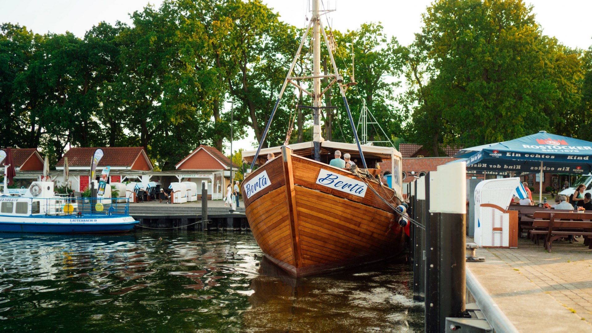 Ein hölzerner Kutter mit dem Namen „Berta“ liegt im Hafen von Lauterbach auf Rügen. Im Hintergrund sind kleine rote Gebäude, Strandkörbe und Sitzbereiche unter Sonnenschirmen sichtbar. Umgeben von ruhigem Wasser und grünen Bäumen bietet die Szenerie eine idyllische Hafenatmosphäre.