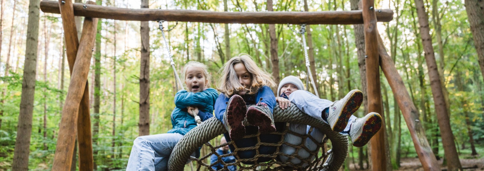 Drei Kinder sitzen gemeinsam auf einer Nestschaukel aus Seilen im Wald, während sie ausgelassen schwingen.