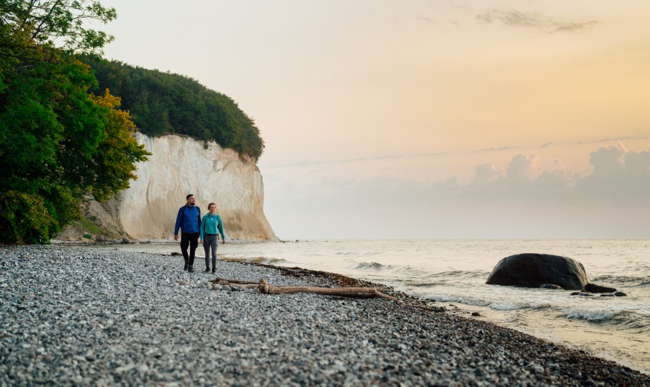 Ein Paar spaziert am Fuße der berühmten Kreidefelsen auf der Insel Rügen entlang, während der Sonnenuntergang den Himmel in sanften Farben erstrahlen lässt.