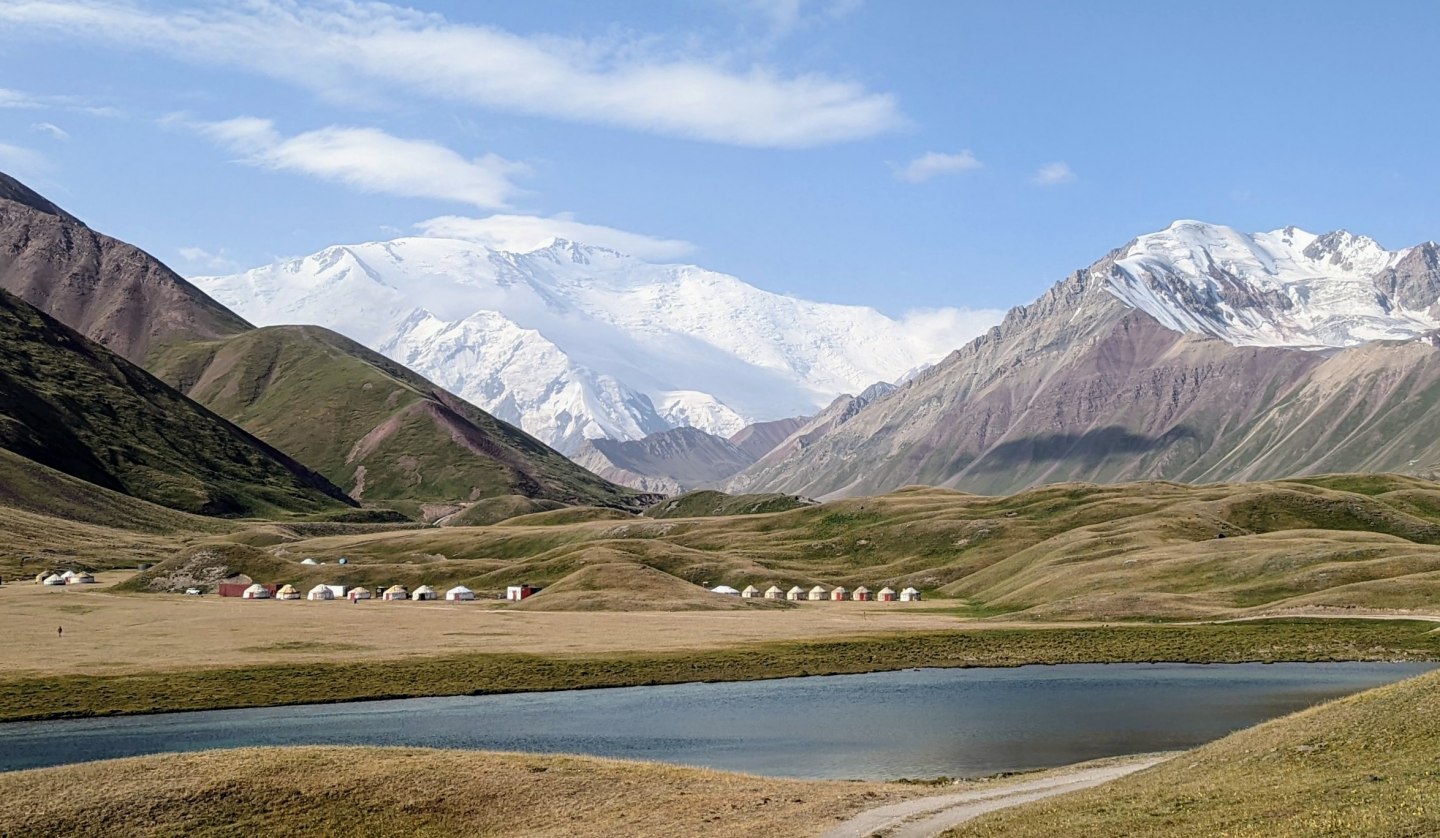 Ein Bild von der Landschaft Kirgistan. Im Hintergrund ist ein Gebirgsmantel, bestehend aus dreu hohen, schneebedeckten Bergen zu sehen.
Davor stehen Hütten von den dortlebenden Menschen und ein Gewässer befindet sich vor ihnen., © Katrin Fischbeck