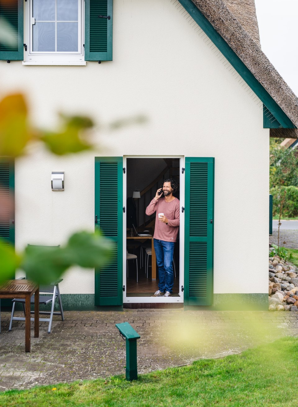 Ein Mann telefoniert und trinkt Kaffee in der Tür eines Ferienhauses mit grünen Fensterläden auf der Insel Poel.