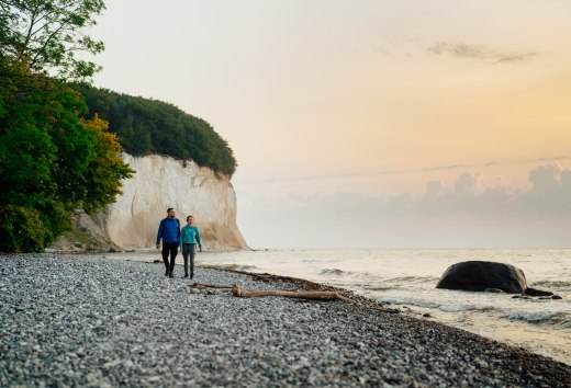 Ein Paar spaziert am Fuße der berühmten Kreidefelsen auf der Insel Rügen entlang, während der Sonnenuntergang den Himmel in sanften Farben erstrahlen lässt.