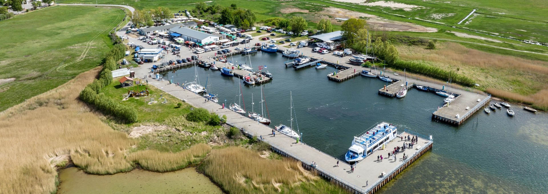 Luftaufnahme des Hafens in Thiessow mit Liegeplätzen für Boote und Schiffe, grünen Wiesen im Hintergrund und der Ostsee unter klarem Himmel.