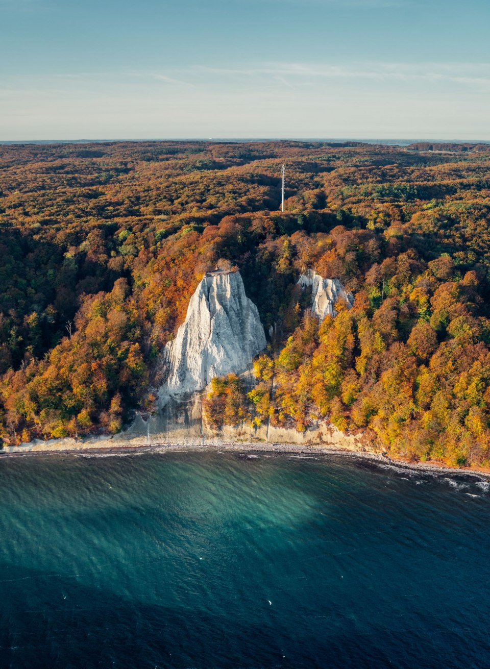 Die imposanten Kreidefelsen im Nationalpark Jasmund leuchten in der morgendlichen Herbstsonne, umgeben von einem Meer aus bunt gefärbtem Laub und der tiefblauen Ostsee – ein atemberaubender Anblick in der goldenen Jahreszeit.