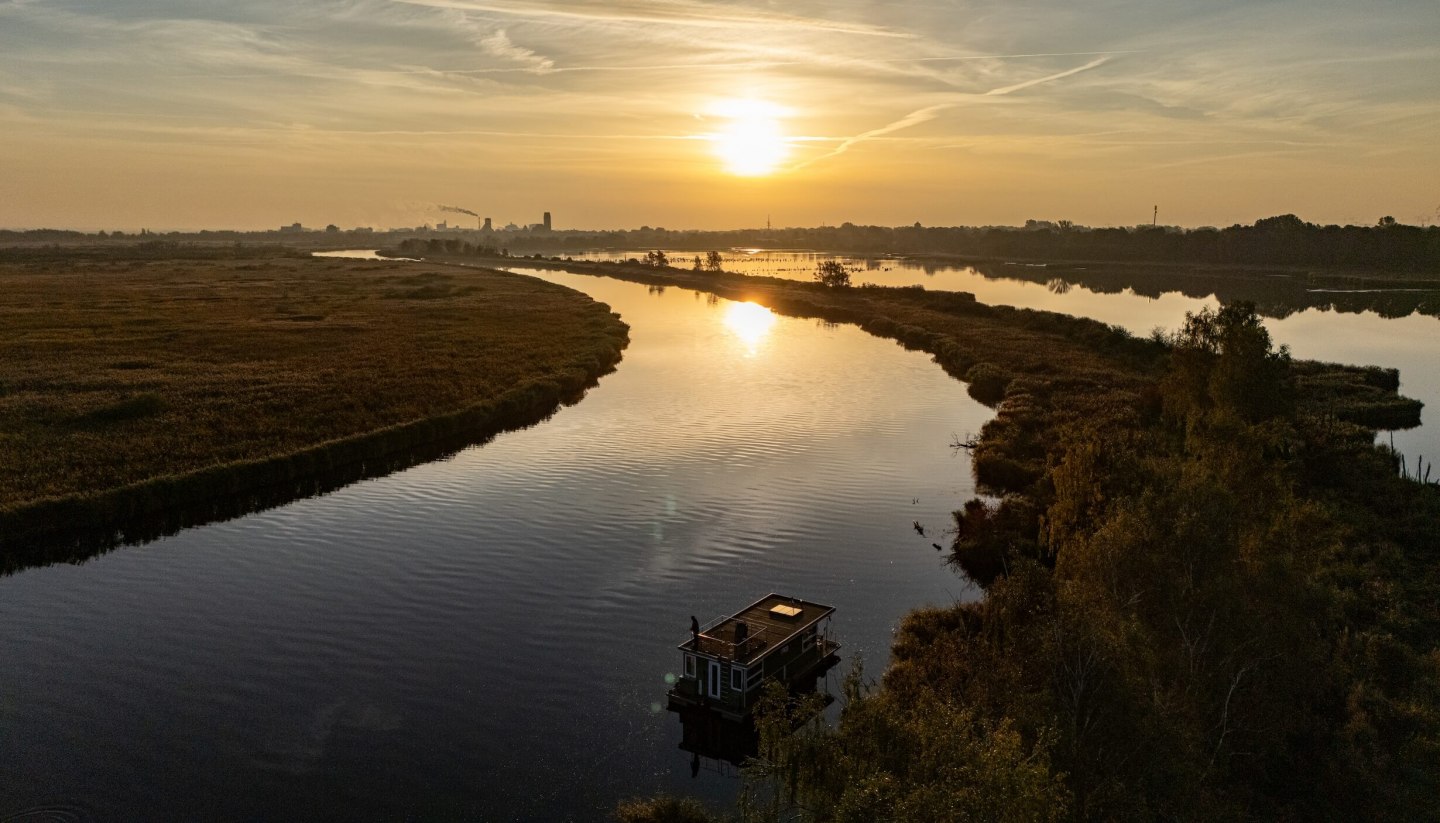 Hausboot auf der Peene bei Sonnenuntergang, umgeben von ruhiger Flusslandschaft und goldenem Licht am Horizont.