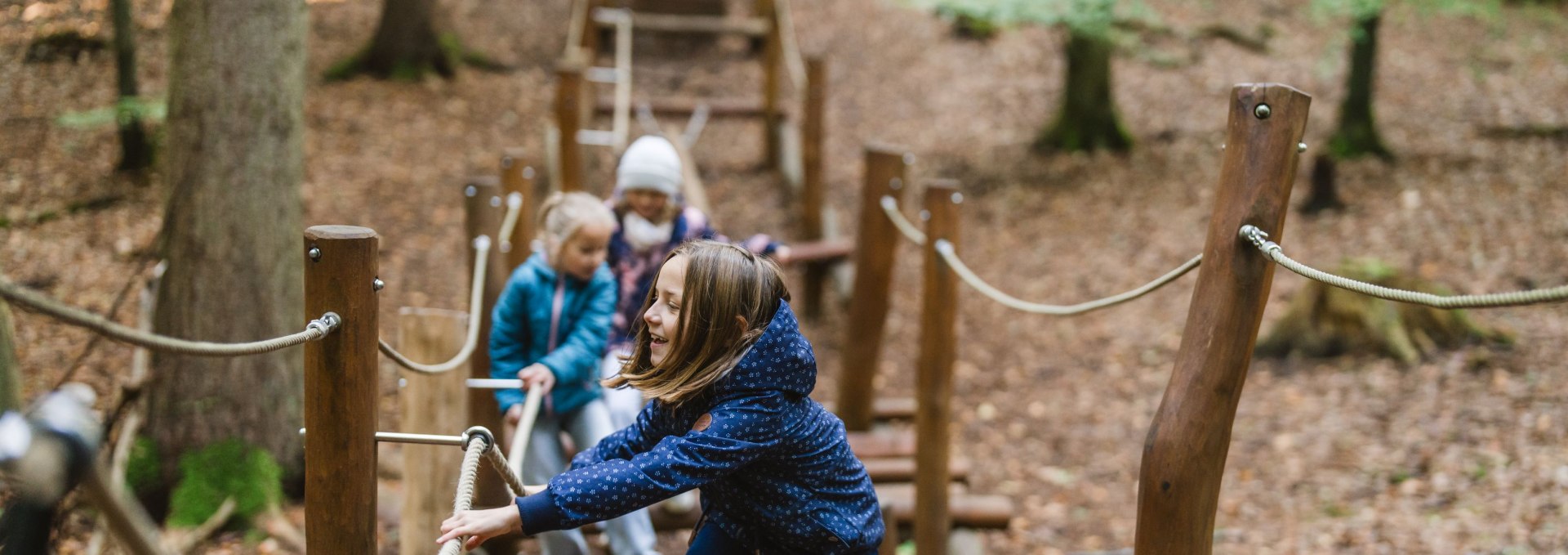 Ein Kind balanciert über einen schmalen Balken auf einem Waldspielplatz, während zwei weitere Kinder im Hintergrund den Parcours entlanglaufen.