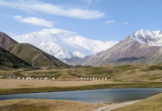 Ein Bild von der Landschaft Kirgistan. Im Hintergrund ist ein Gebirgsmantel, bestehend aus dreu hohen, schneebedeckten Bergen zu sehen.
Davor stehen Hütten von den dortlebenden Menschen und ein Gewässer befindet sich vor ihnen., © Katrin Fischbeck