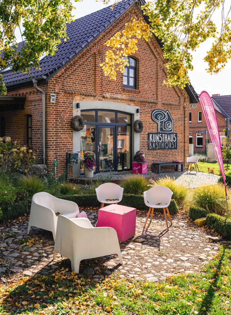 Das Kunsthaus Basthorst in einem herbstlichen Garten, umgeben von gelben und grünen Blättern, mit Sitzgelegenheiten im Außenbereich.