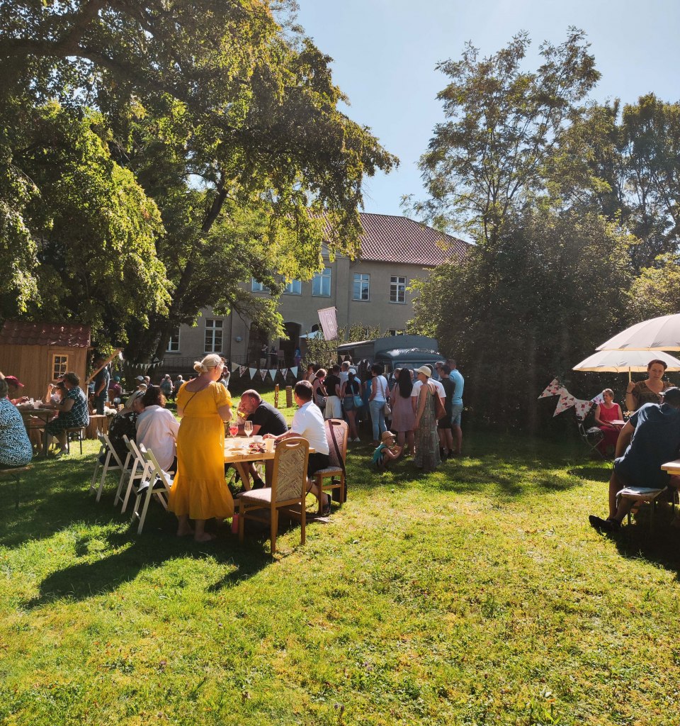 Die MitsommerRemise im Gutspark Klocksin mit Speisen und Getränken. Menschen sitzen an Tischen im Park hinter dem Gutshaus.