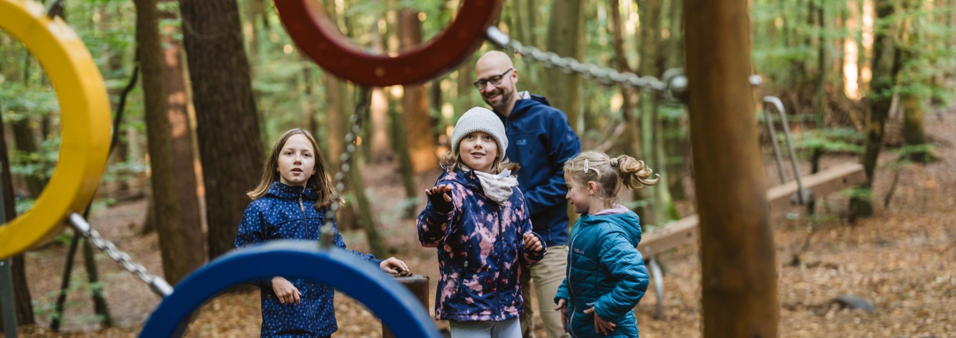 Eine Gruppe von Kindern und ein Erwachsener stehen zwischen bunten Kletterringen aus Metall auf einem Waldspielplatz.