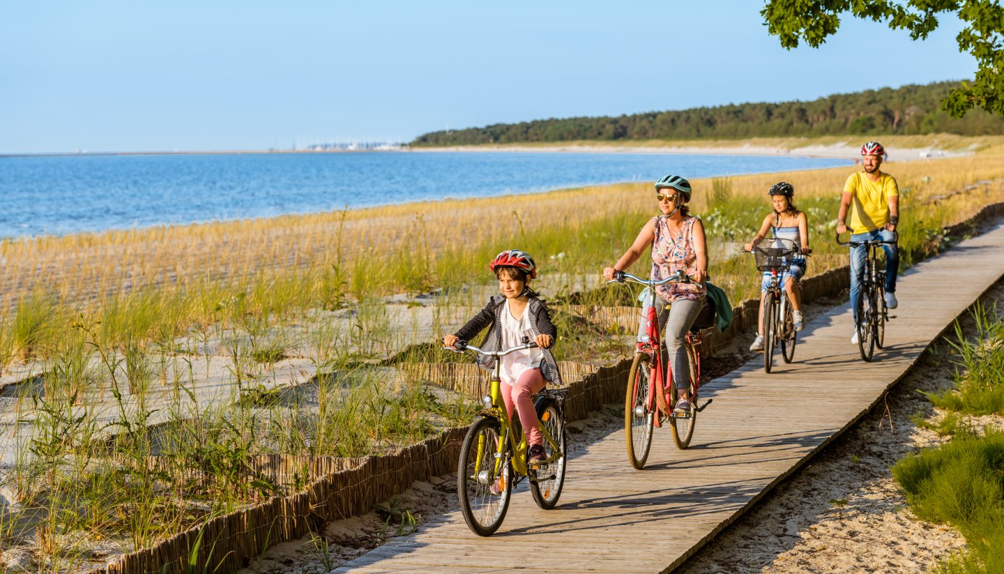 Eine vierköpfige Familie fährt mit dem Fahrrad auf dem Radweg am Strand entlang.