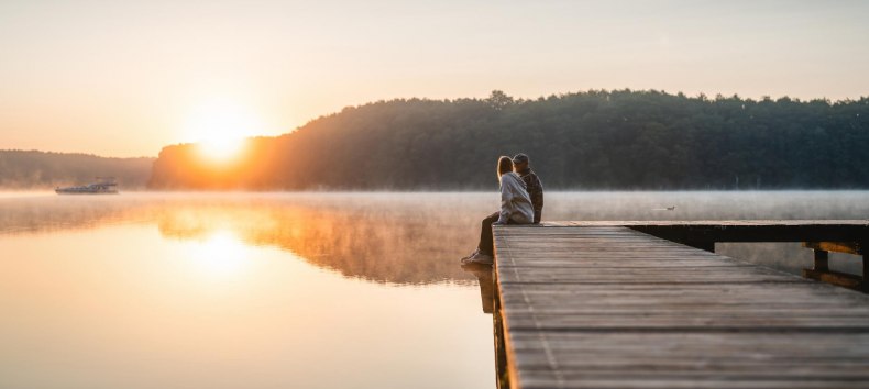 Ein Paar sitzt auf einem Steg zum Sonnenuntergang am Mirower See. Im Hintergrund fährt ein Hausboot vorbei.