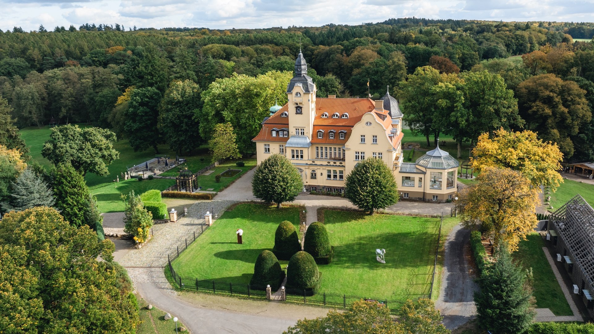 Luftaufnahme des Bernsteinschlosses Wendorf, umgeben von grünen Wiesen und Bäumen unter einem leicht bewölkten Himmel.