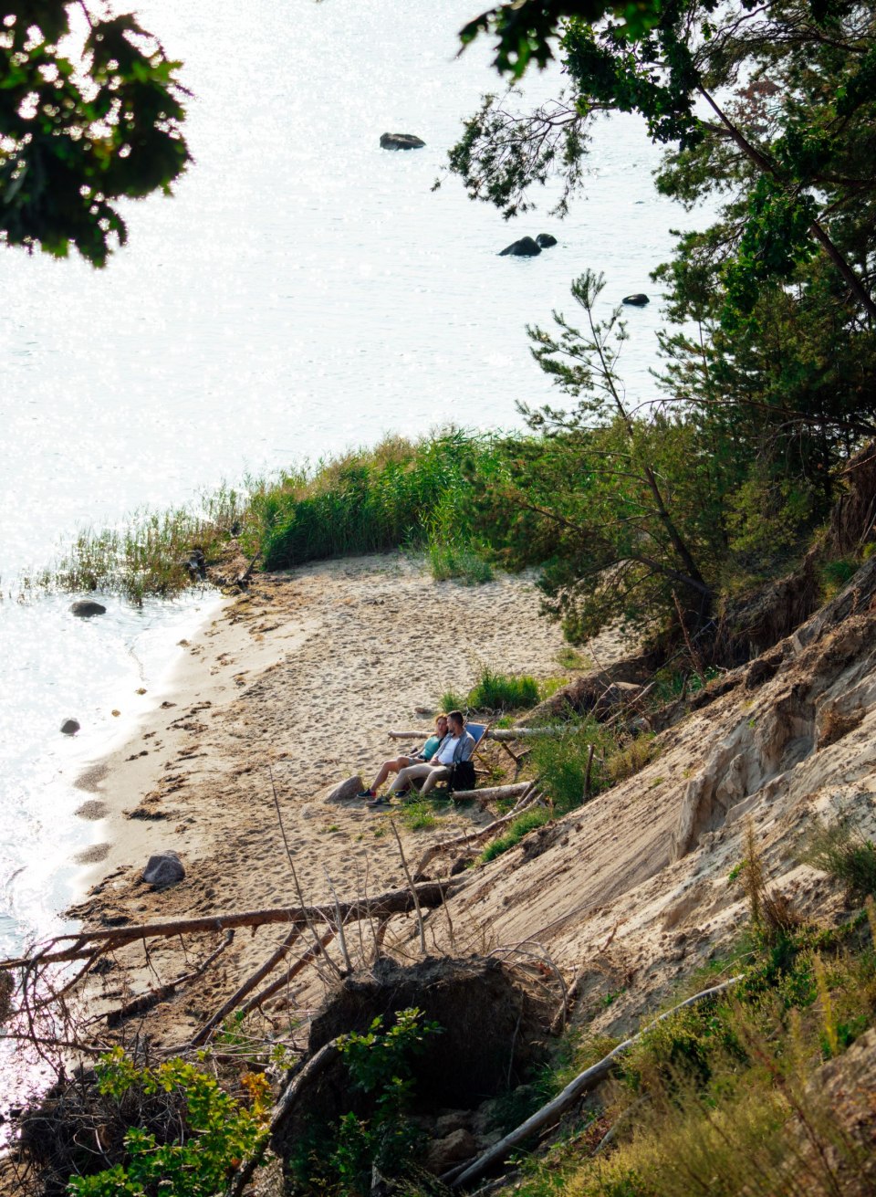 Ein kleiner Sandstrand im Biosphärenreservat Südost-Rügen, umgeben von steilen Uferhängen und dichter Vegetation. Zwei Personen sitzen entspannt auf einer Bank, mit Blick auf das glitzernde Wasser, das von vereinzelten Felsen durchzogen ist. Eine ruhige und abgeschiedene Atmosphäre lädt zum Verweilen ein.