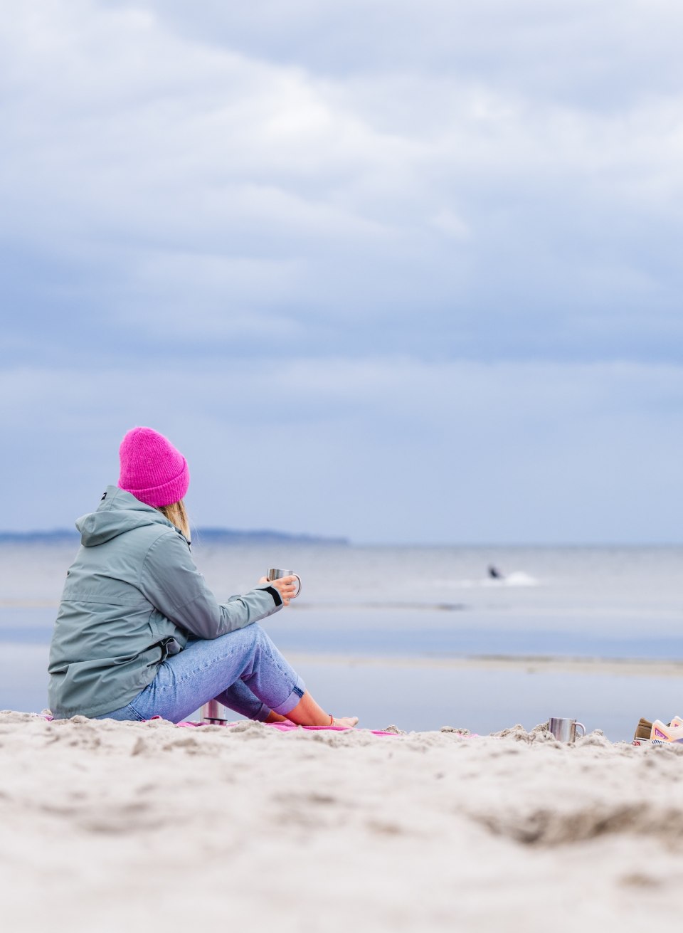  Eine Frau mit pinker Mütze sitzt am Strand von Timmendorf, schaut aufs Meer und hält eine Tasse in der Hand.