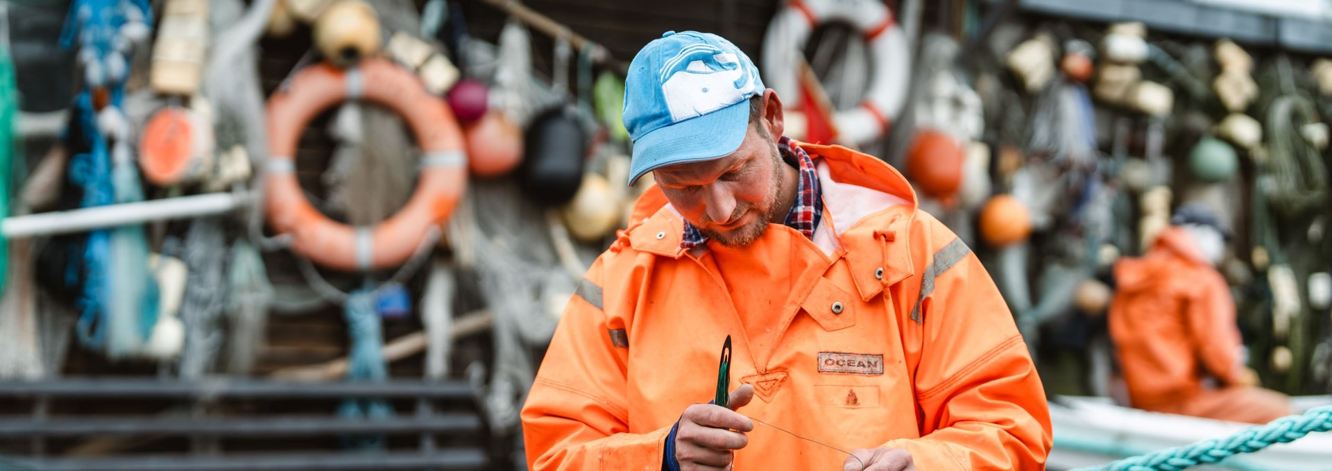 Fischer im Hafen von Gager auf Rügen repariert ein Netz vor einer Hütte mit Rettungsringen und Fischereigeräten, während ein Kollege im Hintergrund sitzt.