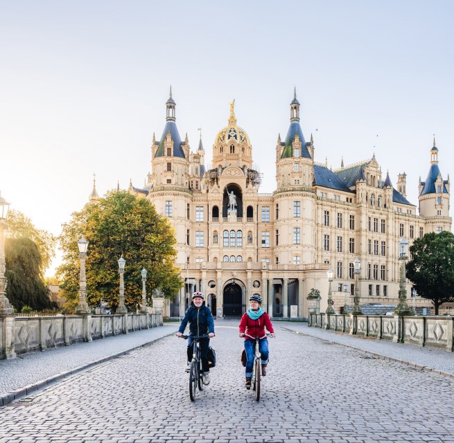 Zwei Radfahrer überqueren eine Brücke vor dem Schweriner Schloss, beleuchtet von der goldenen Morgensonne.