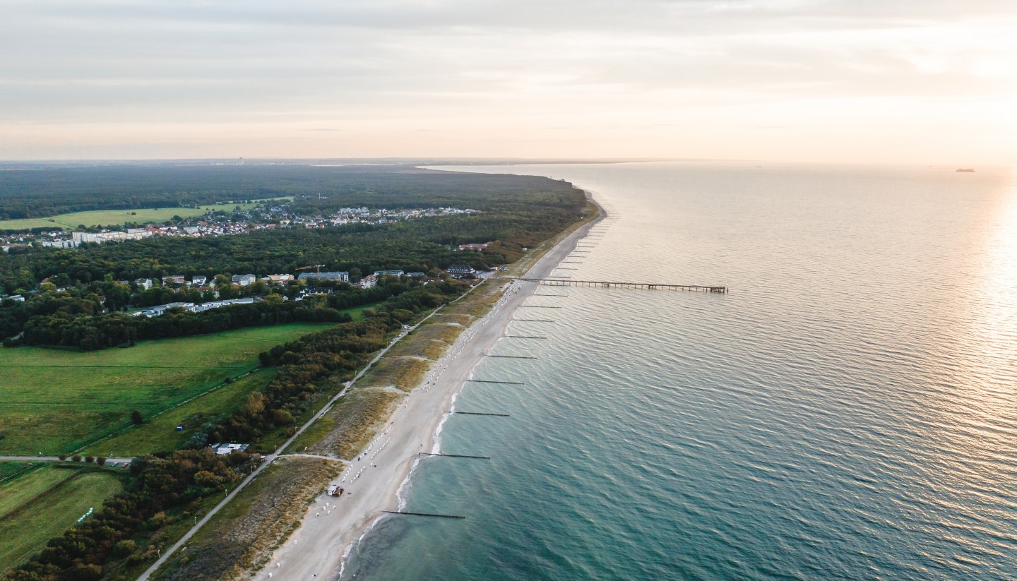 Luftaufnahme der Seebrücke und des Strandes von Graal-Müritz, umgeben von Wald und Wiesen, mit der Ostsee im Licht eines stimmungsvollen Sonnenuntergangs.