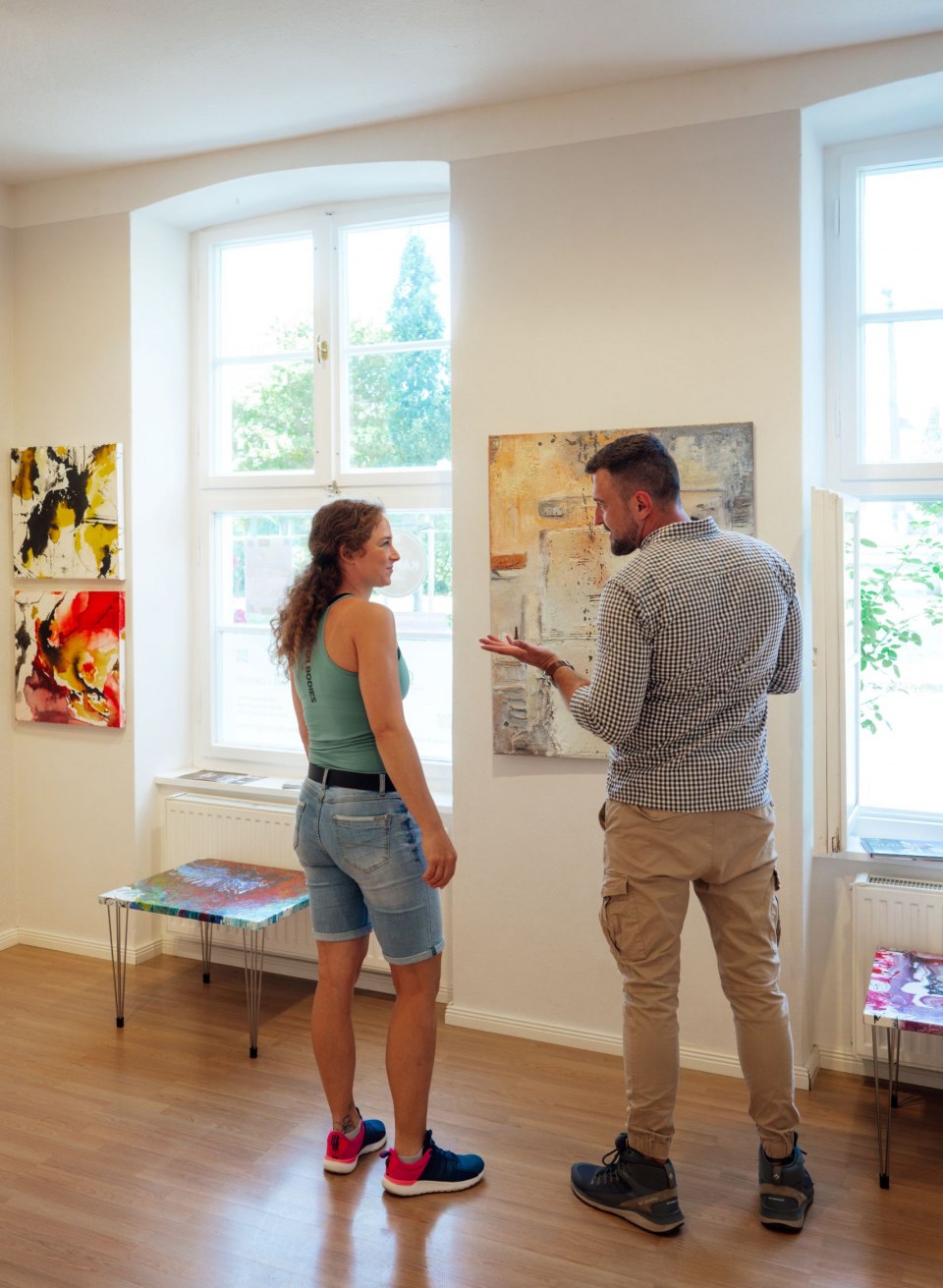 Ein Paar betrachtet abstrakte Gemälde in der Galerie GH Abstrakt in Putbus. An den weißen Wänden hängen farbenfrohe Kunstwerke in verschiedenen Größen. Durch die großen Fenster fällt viel Tageslicht, das die lebhaften Farben der Gemälde betont. Der Raum wirkt hell und einladend.